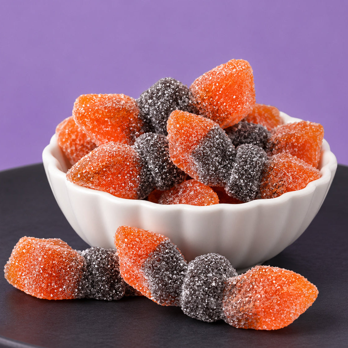 Orange and black sugar-coated sour gummi bats piled in a white scalloped bowl, with two more resting on a dark plate against a purple background