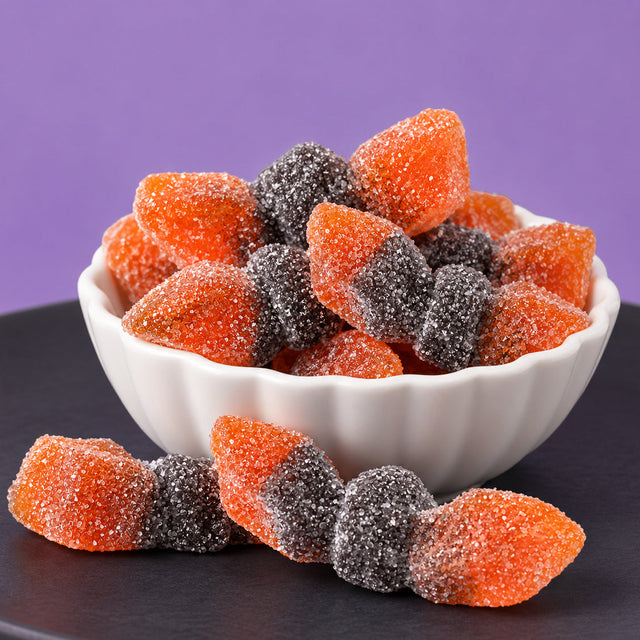 Orange and black sugar-coated sour gummi bats piled in a white scalloped bowl, with two more resting on a dark plate against a purple background