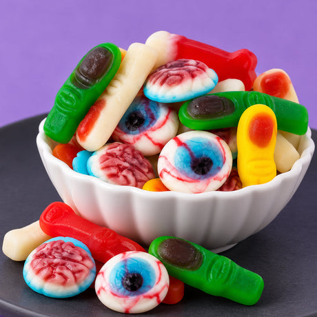 Assorted gummi eyeballs, brains, and fingers piled in a white scalloped bowl, with more pieces on a dark plate against a purple background.
