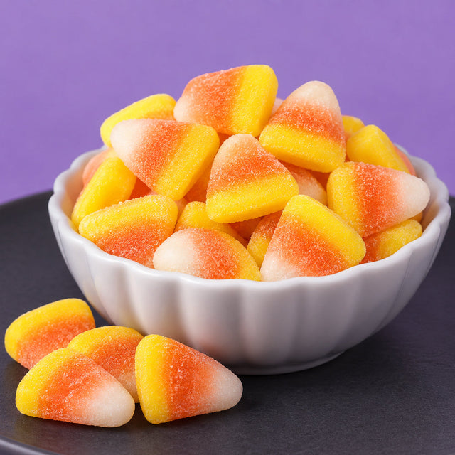 Yellow, orange, and white sugar-coated gummi candy corn piled in a white scalloped bowl, with a few more on a dark plate against a purple background.