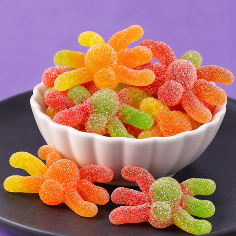 Red, orange, yellow, and green sugar-coated sour gummi spiders piled in a white scalloped bowl, with two more on a dark plate against a purple background.