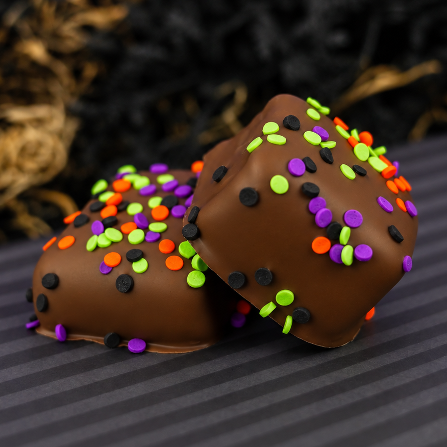 Homemade vanilla marshmallows covered in milk chocolate topped with Halloween confetti sprinkles in orange, green, purple and black, on a striped plate with a purple backdrop. 
