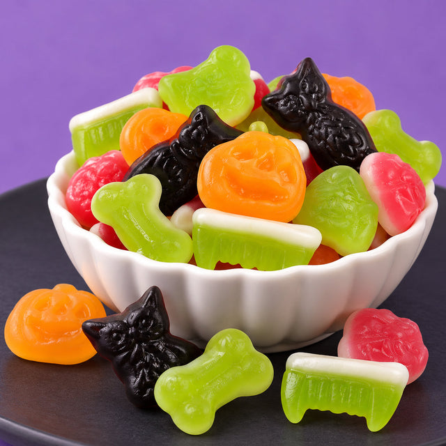 Assorted Halloween gummies including black owls, orange jack-o'-lanterns, green bones and fangs, and pink brains in a white scalloped bowl on a dark plate against a purple background
