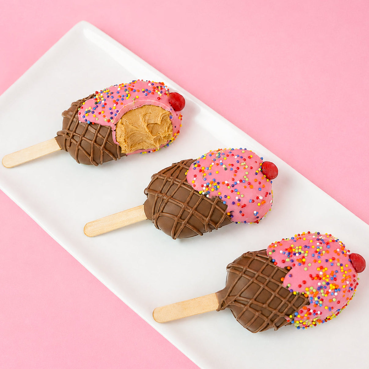 Ice cream milk chocolate peanut butter pop revealing a thick creamy handmade peanut butter ganache center with pink chocolate top and crosshatch milk chocolate cone alongside two whole pops, gourmet summer treat by Nibbles and Bits Scranton PA.