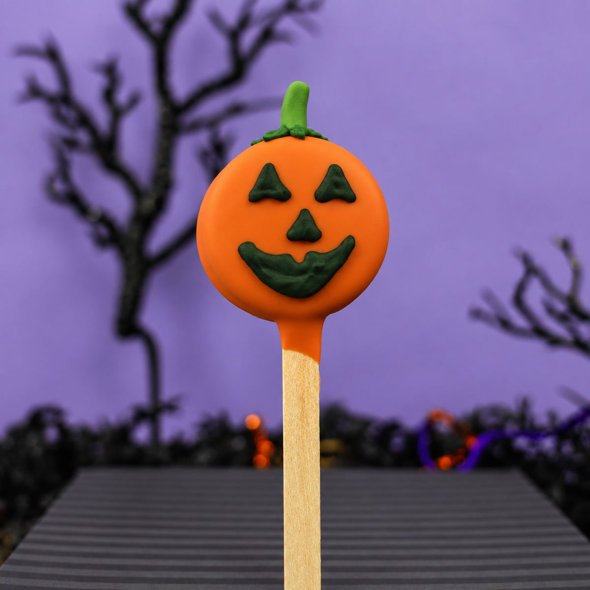 A Halloween-themed pumpkin oreo pop with an orange body, green stem, and carved jack-o'-lantern face, mounted on a wooden stick against a purple background with blurred spooky trees.