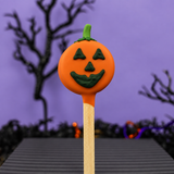 A Halloween-themed pumpkin oreo pop with an orange body, green stem, and carved jack-o'-lantern face, mounted on a wooden stick against a purple background with blurred spooky trees.