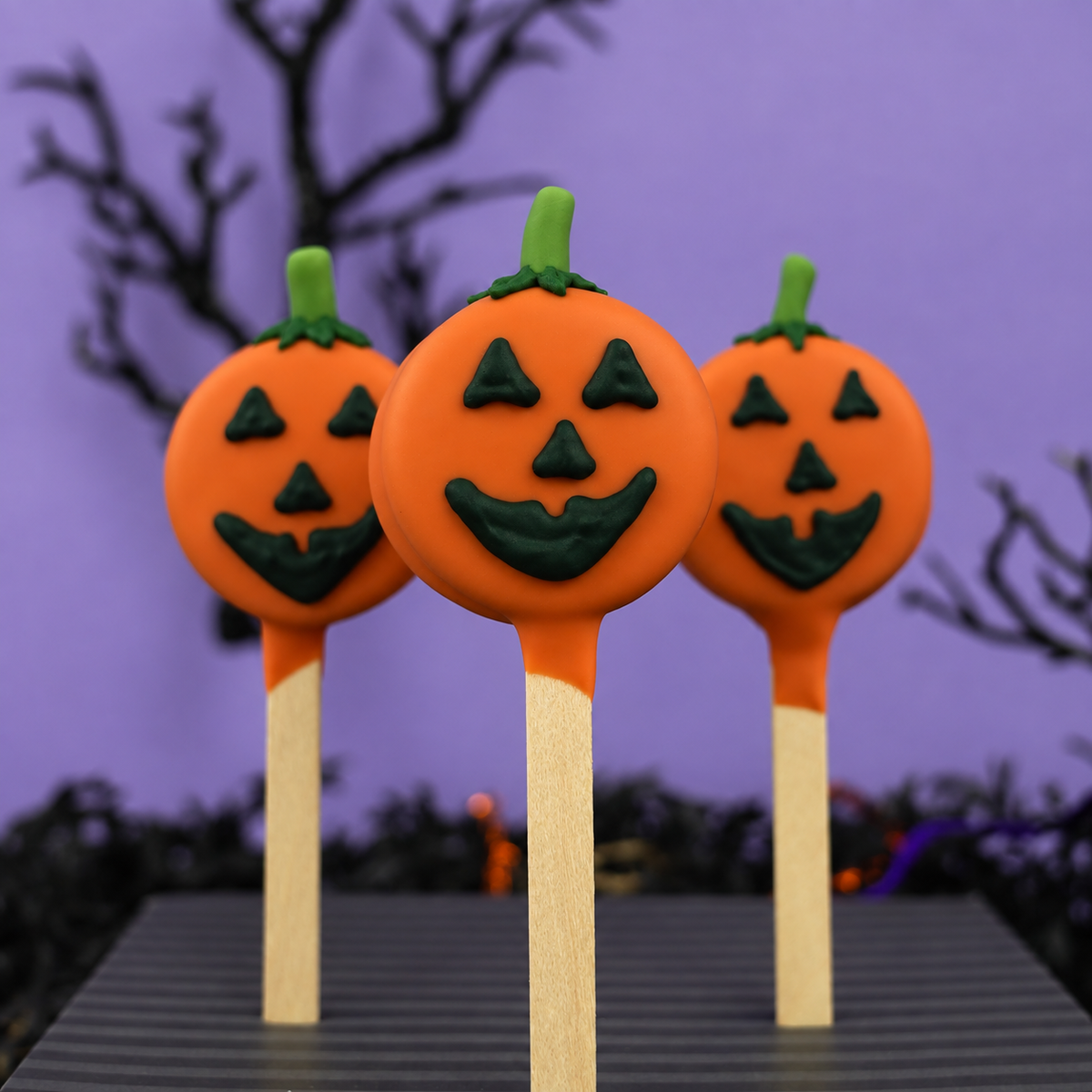 Three Halloween jack-o'-lantern oreo pops with orange and green chocolate decorations on wooden sticks, set against a spooky purple background with silhouettes of trees.