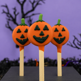 Three Halloween jack-o'-lantern oreo pops with orange and green chocolate decorations on wooden sticks, set against a spooky purple background with silhouettes of trees.