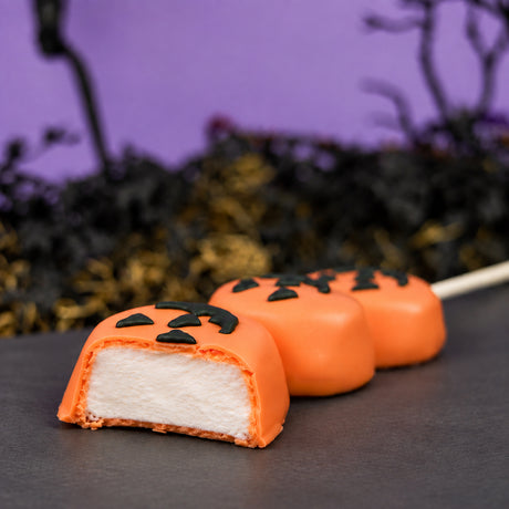 Three homemade vanilla marshmallows on a stick dipped in orange chocolate and hand decorated with jack o lantern faces, cut open to show marshmallow filling. 