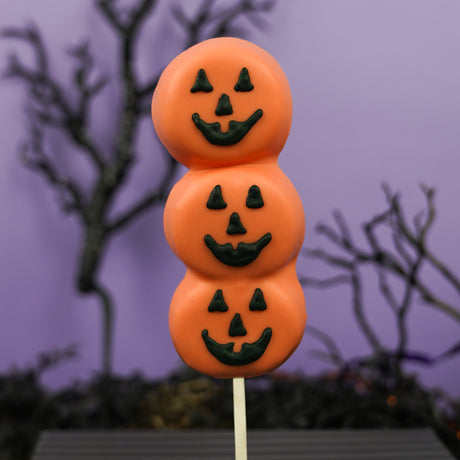 Three handmade vanilla marshmallows on a stick, dipped in orange chocolate and hand painted with three jack o lantern faces in black icing, on a purple halloween background. 