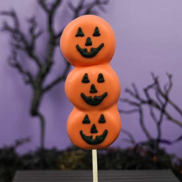 Three handmade vanilla marshmallows on a stick, dipped in orange chocolate and hand painted with three jack o lantern faces in black icing, on a purple halloween background. 