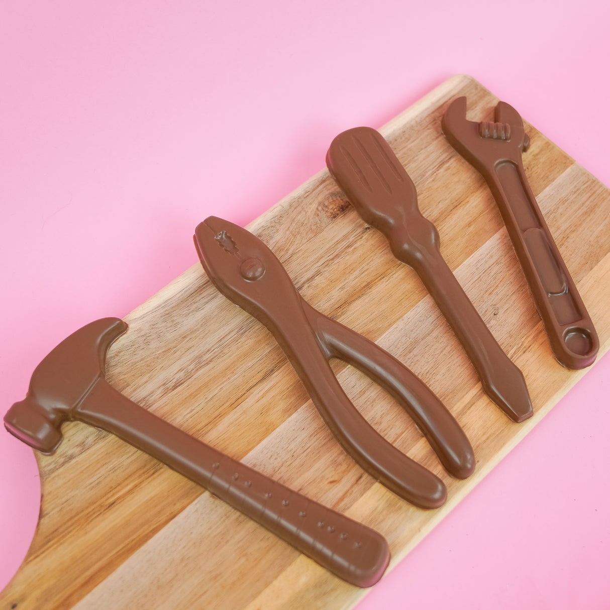 Four large solid milk chocolate tools including a hammer pliers screwdriver and wrench displayed on a rustic wood board with pink background, handmade Father's Day gift set by Nibbles and Bits Scranton PA.