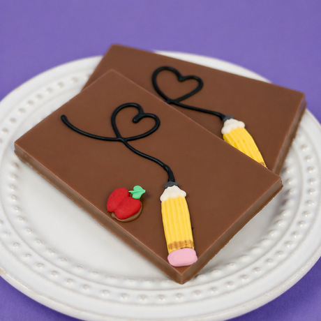 Solid milk chocolate bar hand-decorated with two yellow fondant pencils, a red apple, and a flowing heart design on a white scalloped plate, teacher appreciation gift by Nibbles & Bits.