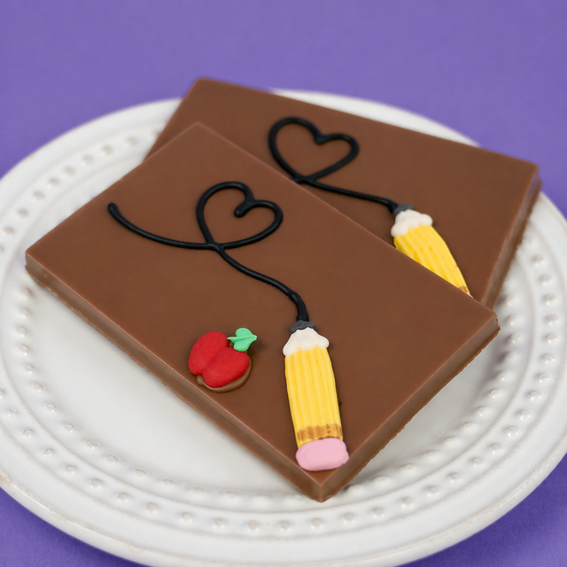 Solid milk chocolate bar hand-decorated with two yellow fondant pencils, a red apple, and a flowing heart design on a white scalloped plate, teacher appreciation gift by Nibbles & Bits.