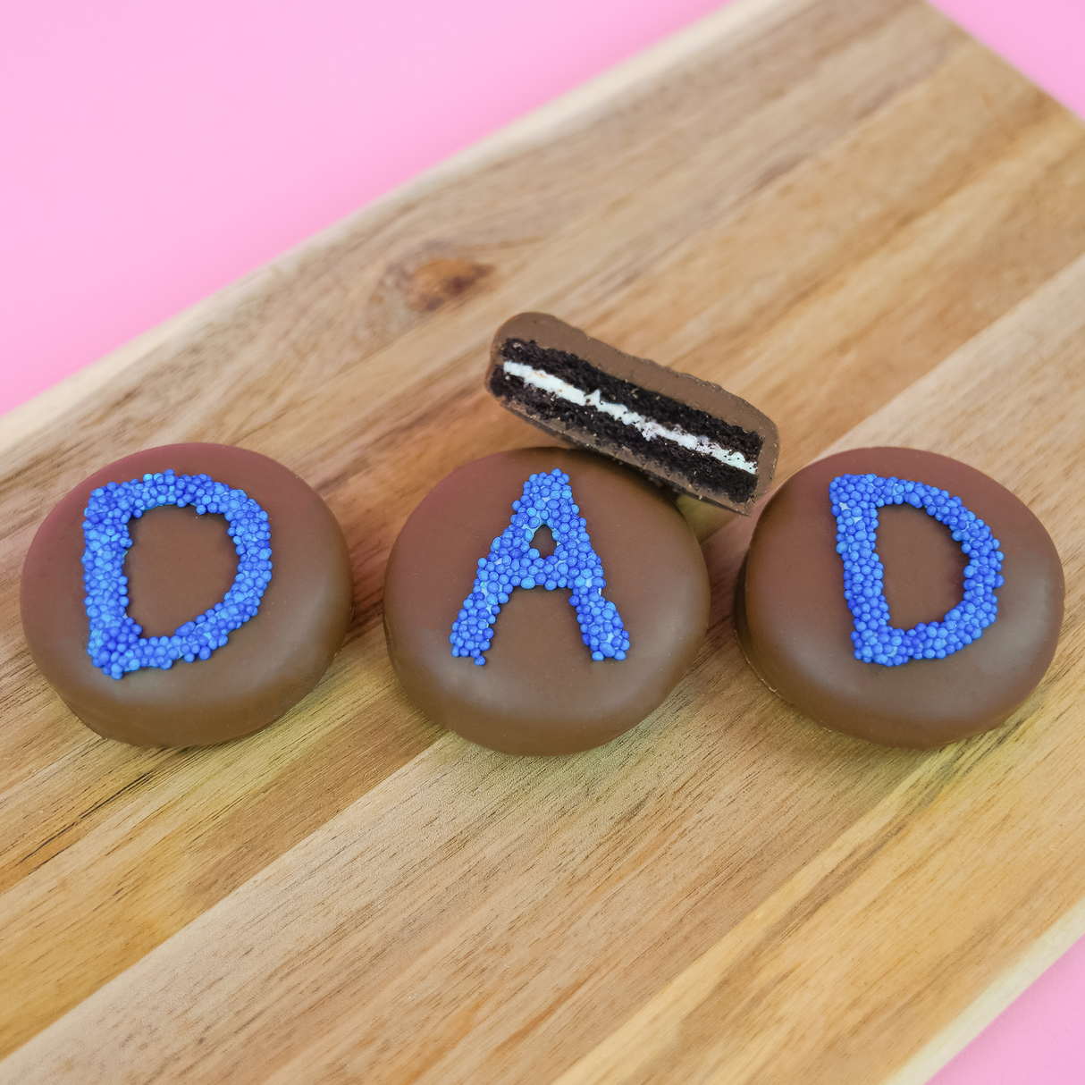 Three milk chocolate DAD Oreos on a wood board with one showing a cross-section of dark chocolate cookie and white cream filling, handcrafted Father's Day chocolate gift by Nibbles and Bits Scranton PA.