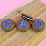 Three milk chocolate DAD Oreos on a wood board with one showing a cross-section of dark chocolate cookie and white cream filling, handcrafted Father's Day chocolate gift by Nibbles and Bits Scranton PA.