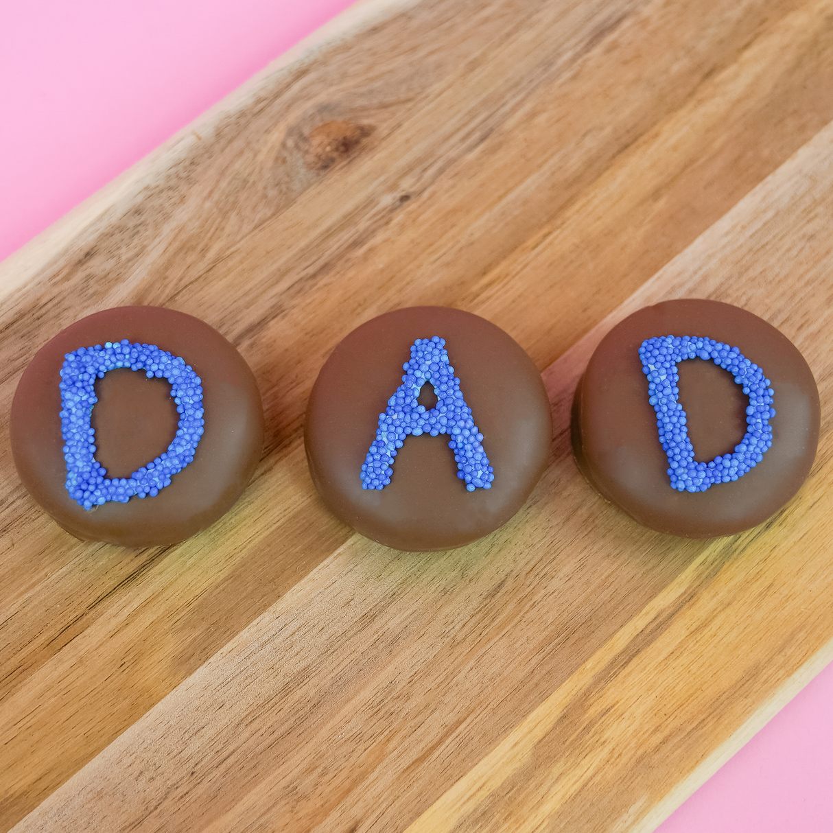 Three milk chocolate DAD Oreos on a wood board with one showing a cross-section of dark chocolate cookie and white cream filling, handcrafted Father's Day chocolate gift by Nibbles and Bits Scranton PA.