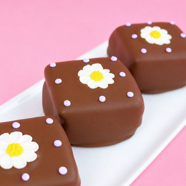 Three milk chocolate daisy s'mores on a white rectangular plate with pink background, each decorated with a white fondant daisy with yellow center and soft lavender icing dots, handmade spring treat by Nibbles and Bits Scranton PA.