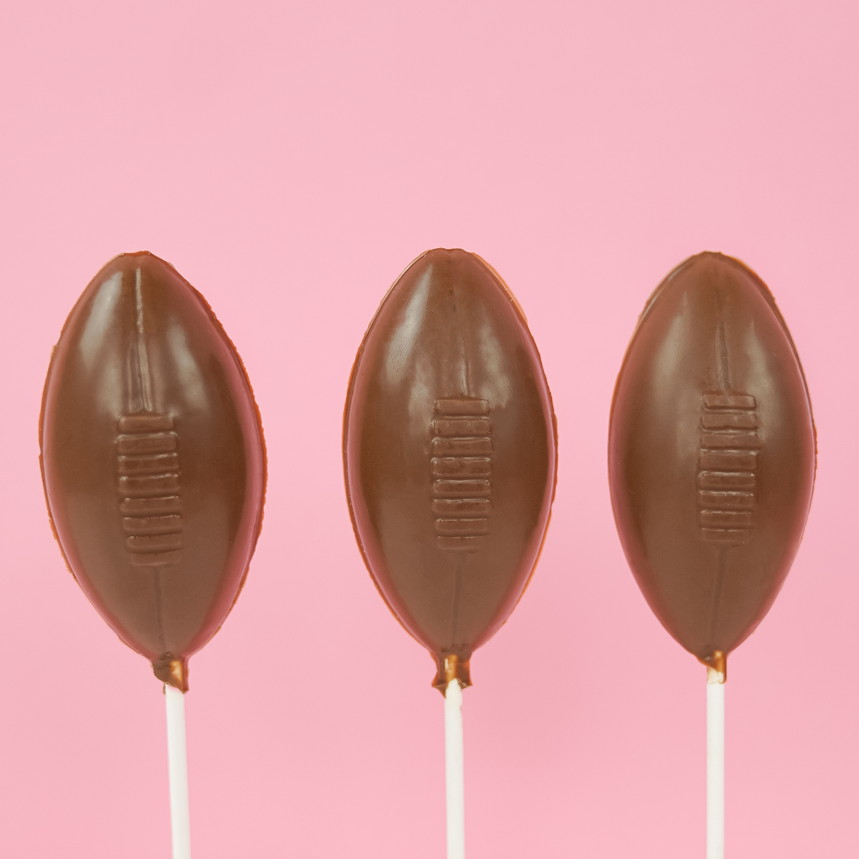 Three solid milk chocolate football shaped lollipops with molded lace stitching detail on white sticks against a pink background, handmade game day treat by Nibbles and Bits Scranton PA.