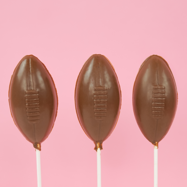 Three solid milk chocolate football shaped lollipops with molded lace stitching detail on white sticks against a pink background, handmade game day treat by Nibbles and Bits Scranton PA.