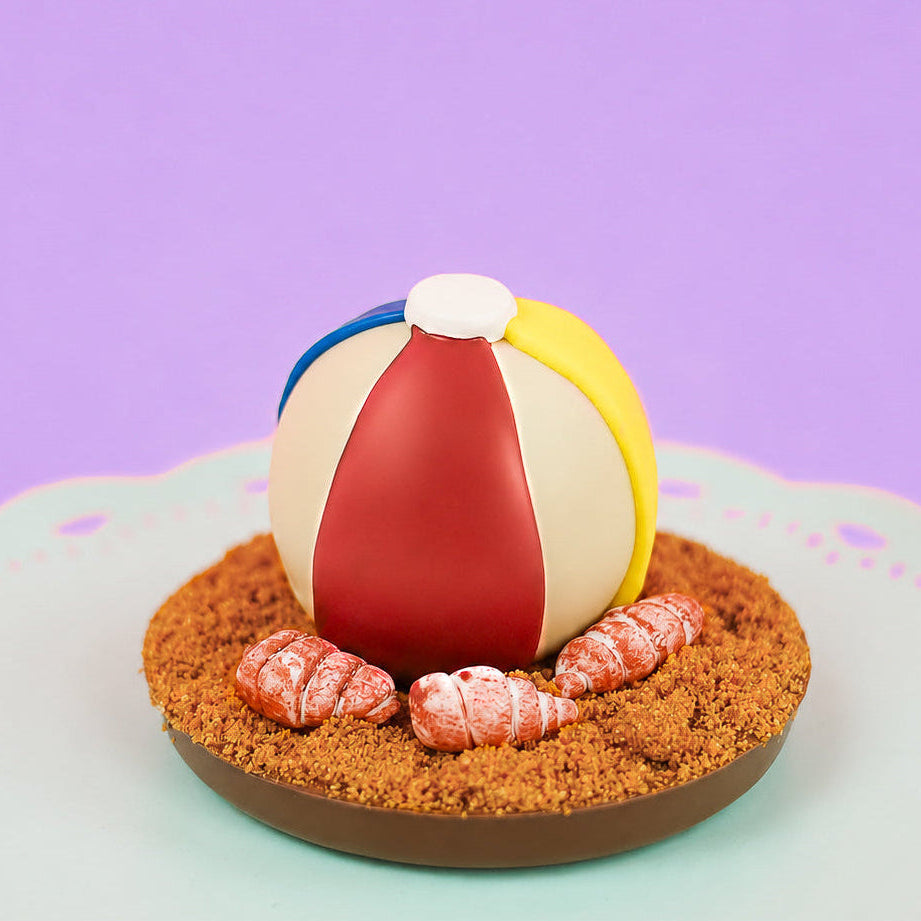 Milk chocolate ganache truffle decorated as a colorful beach ball with yellow, blue and red royal icing stripes resting on a milk chocolate disk covered in crumbled Biscoff cookie sand with hand-painted fondant seashells on a mint green cake stand with purple background, handmade summer gift by Nibbles and Bits Scranton PA.