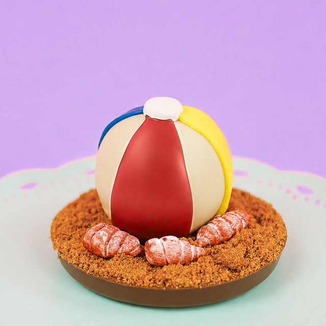 Milk chocolate ganache truffle decorated as a colorful beach ball with yellow, blue and red royal icing stripes resting on a milk chocolate disk covered in crumbled Biscoff cookie sand with hand-painted fondant seashells on a mint green cake stand with purple background, handmade summer gift by Nibbles and Bits Scranton PA.