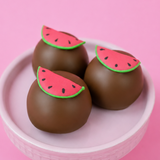 Three milk chocolate ganache watermelon truffles in a pink bowl with pink background, each topped with an adorable fondant watermelon slice with green rind red flesh and black seeds, handmade summer gift by Nibbles and Bits Scranton PA.