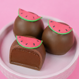 A milk chocolate ganache watermelon truffle revealing a rich creamy milk chocolate ganache center alongside two whole decorated truffles in a pink bowl, handcrafted summer treat by Nibbles and Bits Scranton PA.