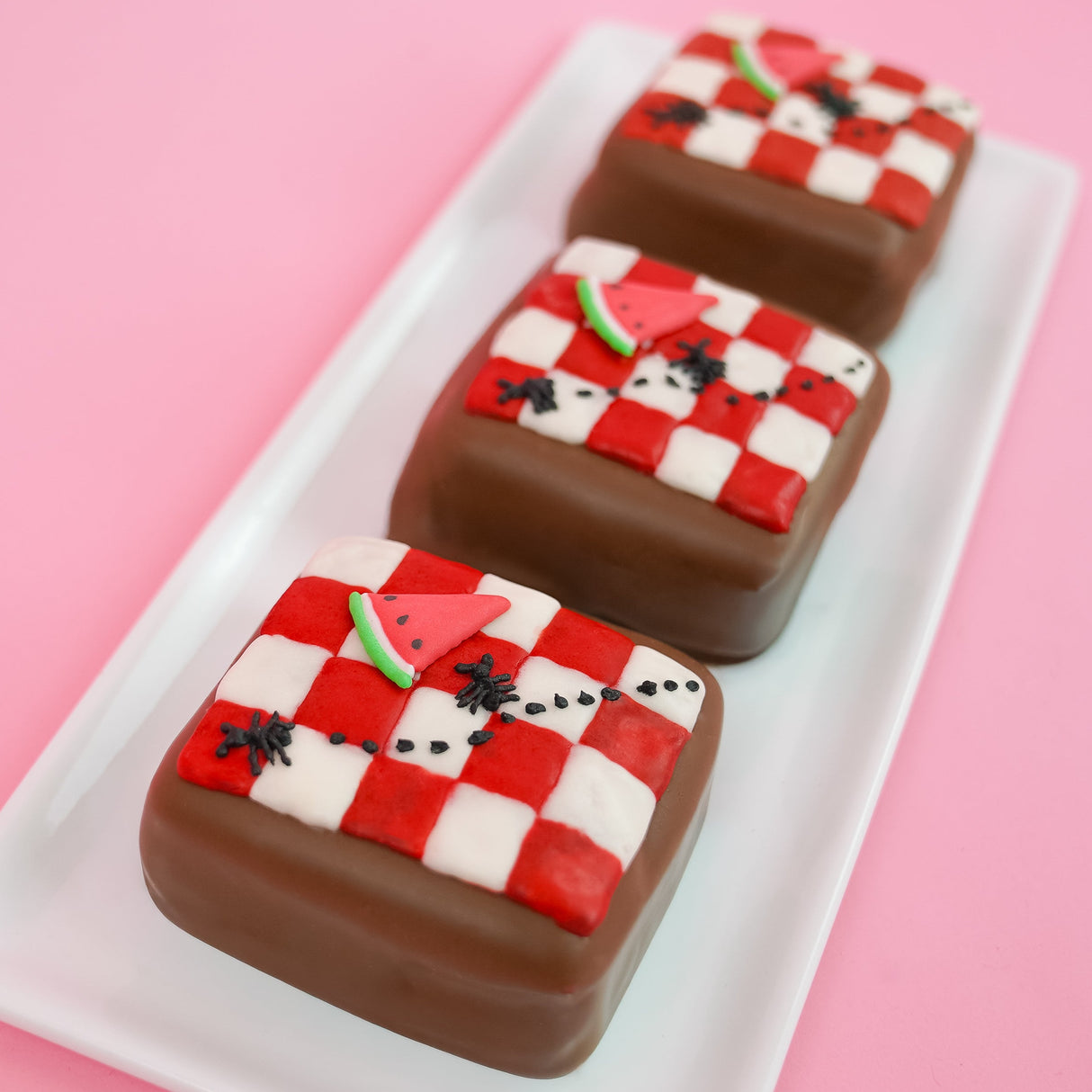 Three milk chocolate s'mores on a white rectangular plate with pink background, each decorated with a hand-made red and white checkered icing picnic blanket with a tiny watermelon slice and ant details, handmade summer treat by Nibbles and Bits Scranton PA.