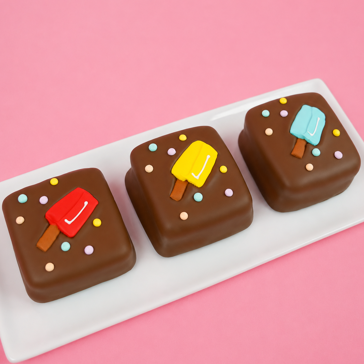 Three milk chocolate popsicle s'mores on a white rectangular plate decorated with red, yellow and blue fondant popsicle decals and colorful pastel dots on a pink background, handmade summer treat by Nibbles and Bits Scranton PA.