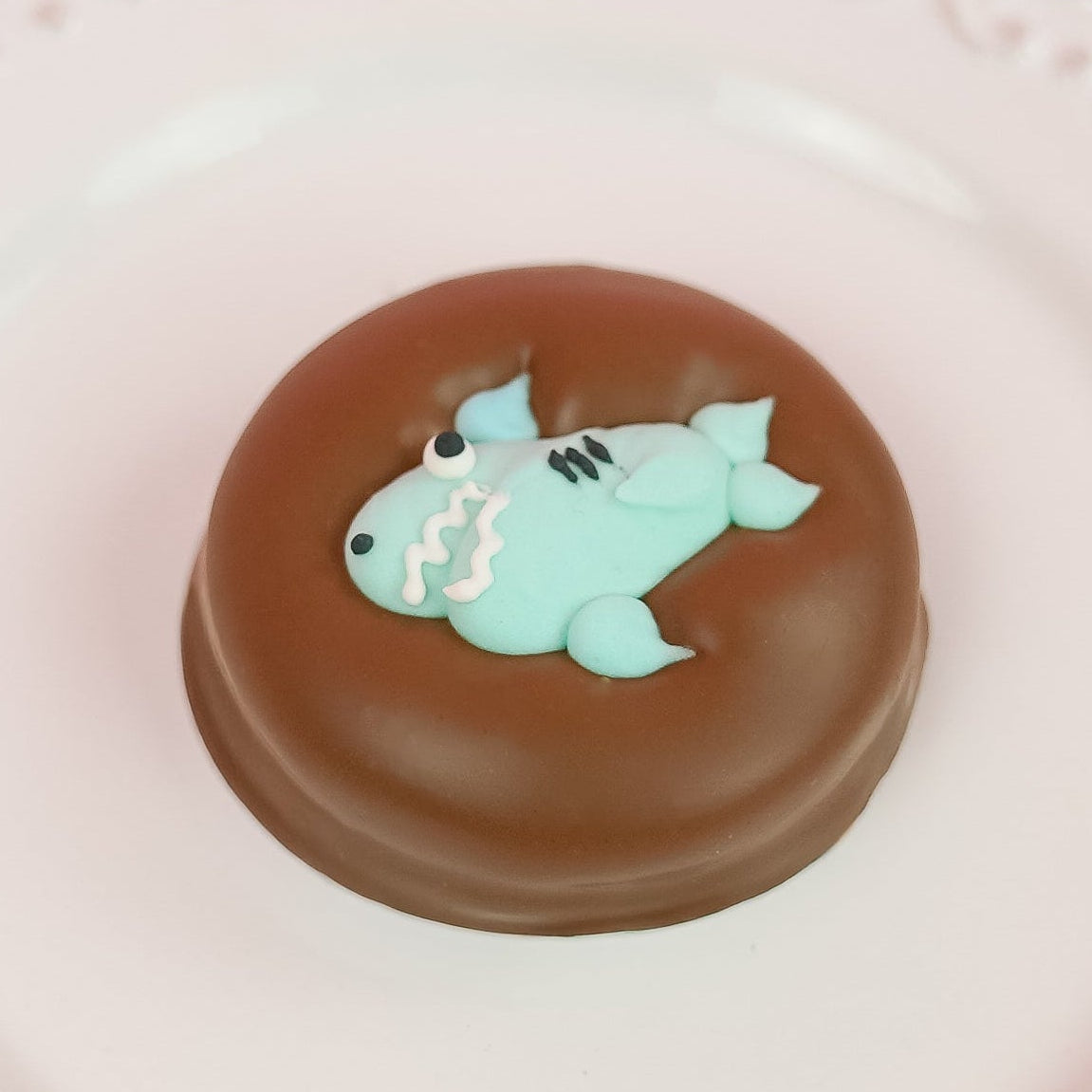 Single milk chocolate covered Oreo decorated with an adorable light blue fondant shark with toothy grin fins and tail on a white decorative plate with a blue background, handmade ocean themed gift by Nibbles and Bits Scranton PA.