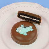 Milk chocolate shark Oreo with dark chocolate cookie layers and white cream filling on a white decorative plate with blue background, handcrafted ocean themed chocolate treat by Nibbles and Bits Scranton PA.
