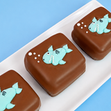 Three milk chocolate shark s'mores on a pink rectangular plate with blue background, each decorated with an adorable teal fondant shark with fins toothy grin and white bubble details, handmade ocean themed treat by Nibbles and Bits Scranton PA.
