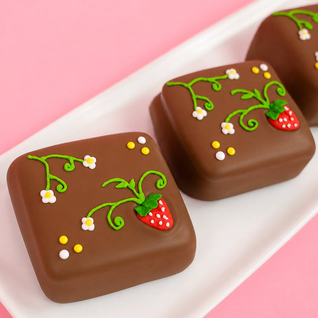 Three milk chocolate strawberry vine s'mores on a white rectangular plate with pink background, each hand-decorated with royal icing green strawberry vines white daisy blooms yellow dot details and a red fondant strawberry with white dots and green leaf cap, handmade spring treat by Nibbles and Bits Scranton PA.