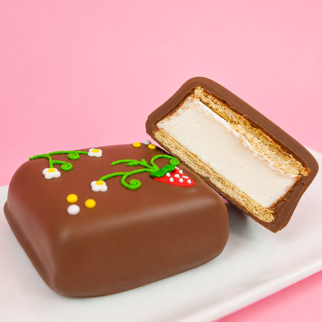 A milk chocolate strawberry vine s'more revealing thick homemade vanilla marshmallow layer and golden graham cracker base alongside a whole decorated s'more on a white plate with pink background, handcrafted spring treat by Nibbles and Bits Scranton PA.