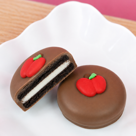 Two chocolate sandwich cookies decorated with red apple shapes and green stems, one whole cookie and one cut open to show the white cream filling.