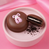 Milk chocolate covered sandwich cookies in a soft pink dish, one decorated with pink ballet slippers icing and one cut in half to reveal a cream filling, styled on a smooth pink background.