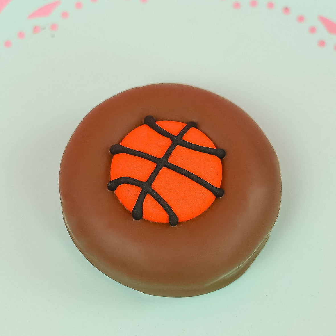 Single milk chocolate covered Oreo decorated with a vibrant orange fondant basketball with dark chocolate seam lines on a mint green decorative plate with pink background, handmade sports gift by Nibbles and Bits Scranton PA.