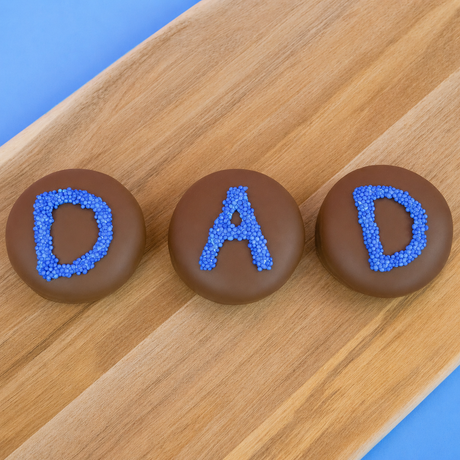 Three milk chocolate DAD Oreos on a wood board with a blue background, handcrafted Father's Day chocolate gift by Nibbles and Bits Scranton PA.
