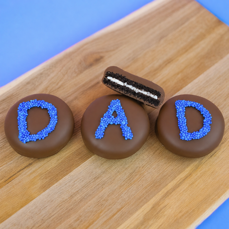 Three milk chocolate DAD Oreos on a wood board with one showing a cross-section of dark chocolate cookie and white cream filling, handcrafted Father's Day chocolate gift by Nibbles and Bits Scranton PA.