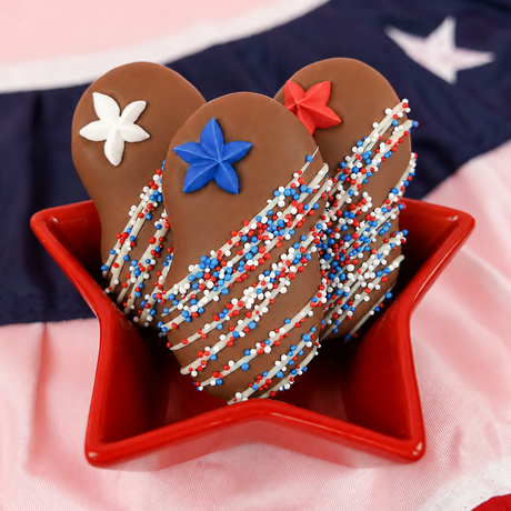 Close-up of milk chocolate Nutter Butter cookie topped with a blue fondant star decal and red, white and blue nonpareils with white chocolate drizzle, patriotic Fourth of July treat by Nibbles and Bits.
