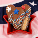 Close-up of milk chocolate Nutter Butter cookie topped with a blue fondant star decal and red, white and blue nonpareils with white chocolate drizzle, patriotic Fourth of July treat by Nibbles and Bits.