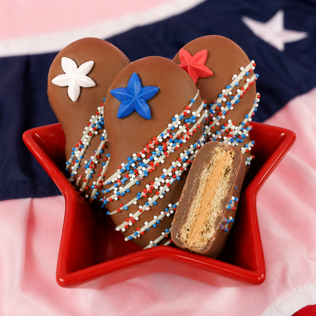 Close-up of milk chocolate Nutter Butter cookie topped with a blue fondant star decal and red, white and blue nonpareils with white chocolate drizzle, patriotic Fourth of July treat by Nibbles and Bits.