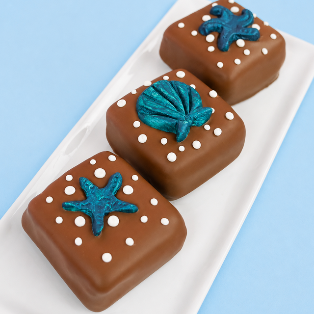 Three milk chocolate sea shell s'mores on a white rectangular plate with blue background, each decorated with a hand-painted metallic teal fondant ocean creature including a starfish a scallop shell and a sea creature surrounded by white bubble dot details, handmade coastal gift by Nibbles and Bits Scranton PA.