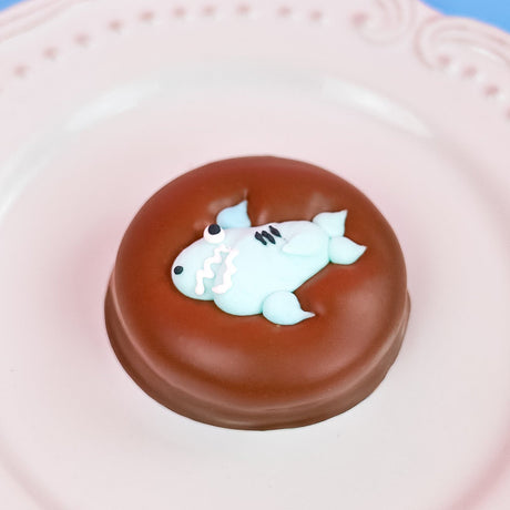 Single milk chocolate covered Oreo decorated with an adorable light blue fondant shark with toothy grin fins and tail on a white decorative plate with a blue background, handmade ocean themed gift by Nibbles and Bits Scranton PA.