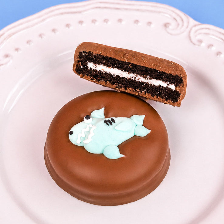 Milk chocolate shark Oreo with dark chocolate cookie layers and white cream filling on a white decorative plate with blue background, handcrafted ocean themed chocolate treat by Nibbles and Bits Scranton PA.