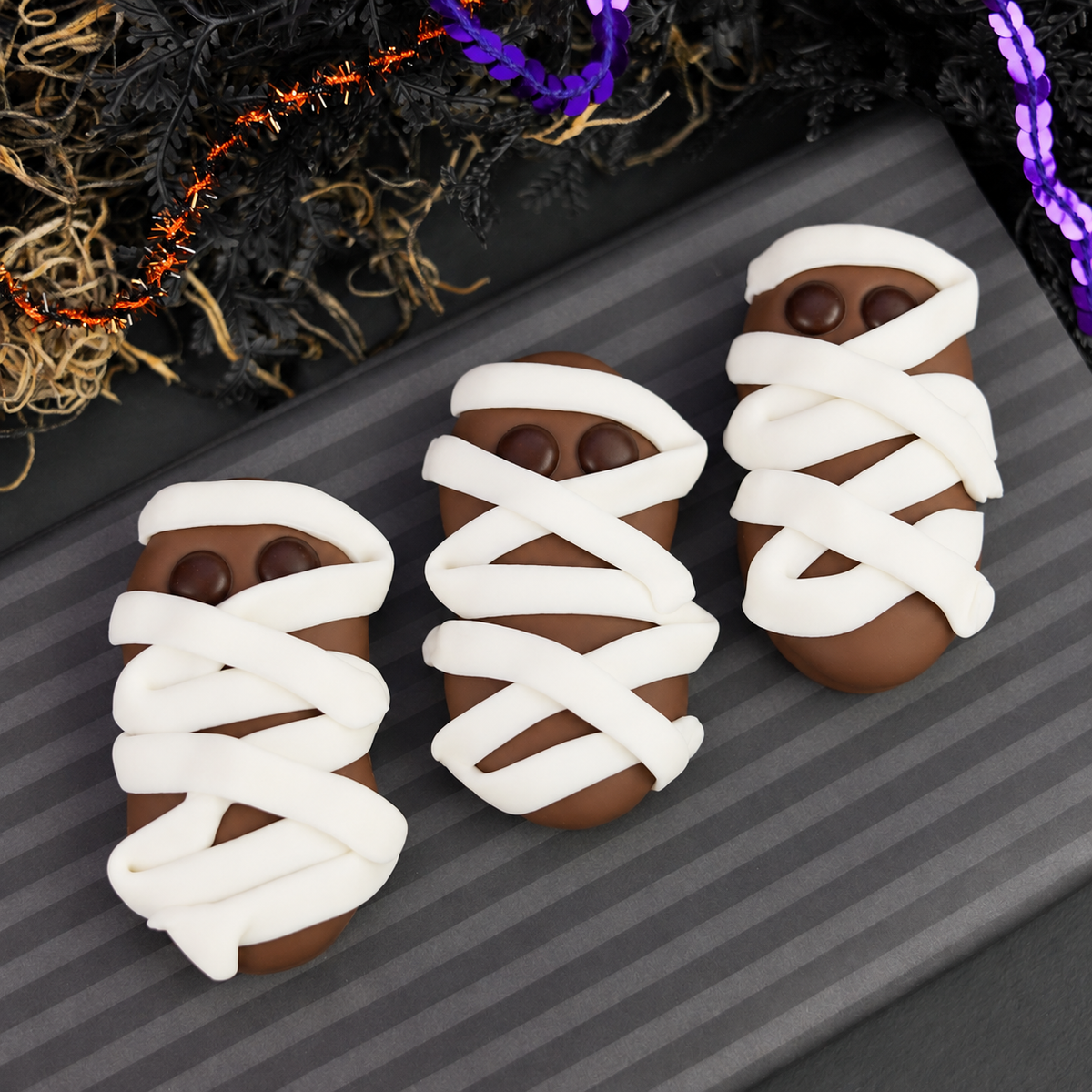Three Halloween mummy nutter butter cookies, wrapped in white icing with small brown eyes, arranged on a dark gray striped board with decorative black and purple garlands in the background.
