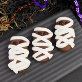 Three Halloween mummy nutter butter cookies, wrapped in white icing with small brown eyes, arranged on a dark gray striped board with decorative black and purple garlands in the background.