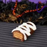 A cut open chocolate-covered nutter butter topped with white icing wrapping, displayed on a dark striped surface with Halloween decorations blurred in the background.