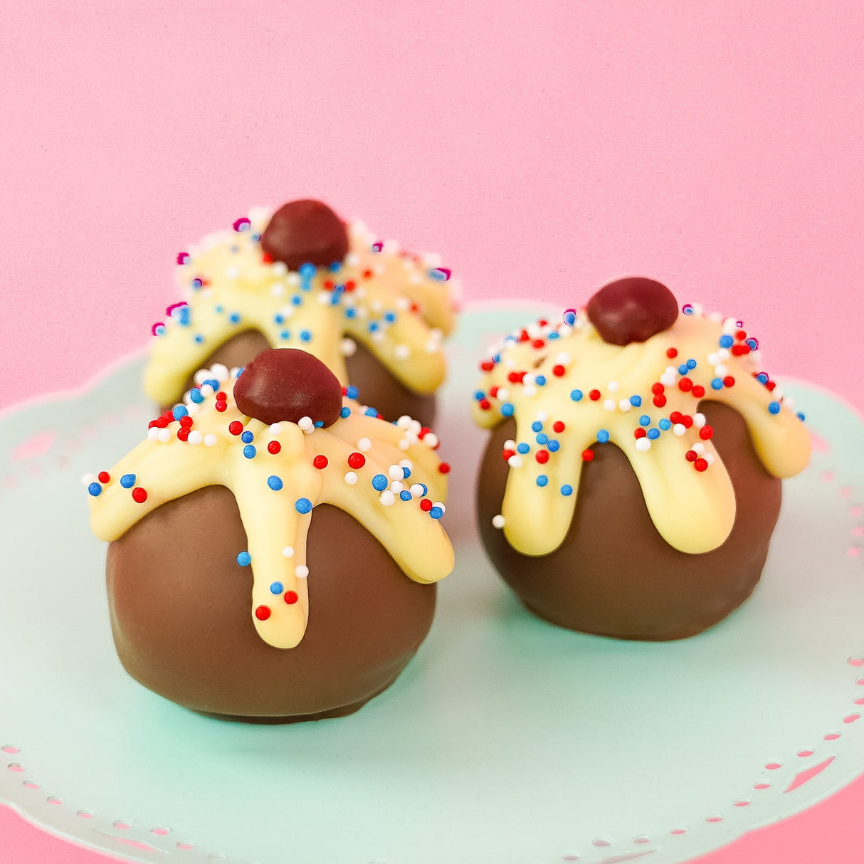 Three patriotic sundae milk chocolate ganache truffles on a mint green cake stand with pink background, each topped with dripping white chocolate, red, white and blue nonpareils and a dark chocolate cherry, handmade Fourth of July gift by Nibbles and Bits Scranton PA.